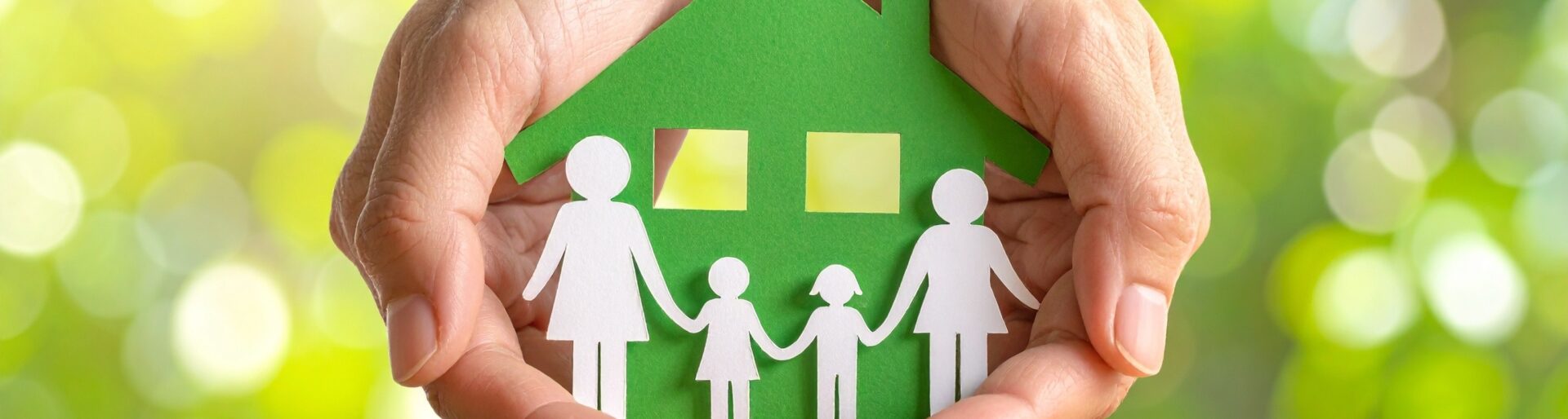 Two hands form a protective roof over a green house icon with a family silhouette on a grassy field, symbolizing eco-friendly living, safety, and community care in a sunlit natural setting.